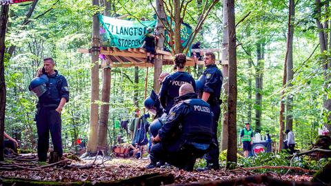 Mehrere Polizeikräfte stehen im Wald vor einer Art Baumplattform mit mit einem Banner auf welchem steht: "Jetzt Langt's Sehring stoppen“.
