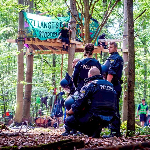 Mehrere Polizeikräfte stehen im Wald vor einer Art Baumplattform mit mit einem Banner auf welchem steht: "Jetzt Langt's Sehring stoppen“.