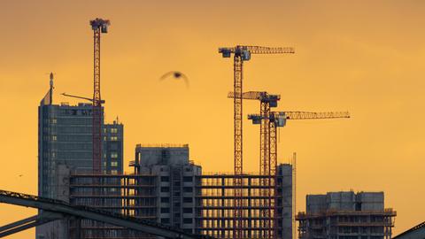 Im Gegenlicht der aufgehenden Sonne zeichnen sich beim Blick von der Flößerbücke in Frankfurt am Main die Hochhäuser und Baukräne der Großbaustelle am Kaiserleikreisel in Offenbach am Main ab.