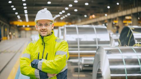 Mann in gelber Sicherheitskleidung mit Helm in Produktionshalle. Im Hintergrund Stahlbündel