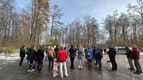 Protest gegen Windräder im Reinhardswald: Eine Gruppe von Menschen, allesamt von hinten fotografiert, steht auf einer freien Fläche vor einem Wald, auf dem Boden etwas Schnee und Eis, unter blauem Himmel, im Hintergrund ist ein Polizeiauto zu erkennen