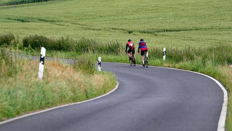 Zwei Rennradfahrer fahren auf einer Landstraße, umgeben von grünen Feldern.