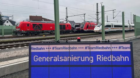 Ein neben Bahngleisen aufgebautes Display mit der Aufschrift "Generalsanierung Riedbahn", daneben ein Mann in Warnweste von hinten, dahinter stehende Züge