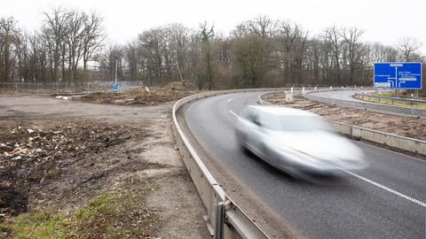 Eine leere Straße führt mit einer Kurve nach rechts. Ein verschwommenes, silbernes Auto bewegt sich schnell auf der Straße. Links ist eine unbebaute Fläche mit Müll zu sehen. Im Hintergrund stehen kahle Bäume. Rechts steht ein blaues Autobahnschild mit den Aufschriften: "F-Bonames", "F-RiederWald", "Bad Vilbe", "Karben-Petterweil", "Friedberg", "F-Praunheim", "Miquelallee", "Nordweststadt", "Bockenheim, Praunheim Nordweststadt".