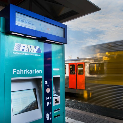 Ein Fahrkartenautomat des RMV in Friedrichsdorf. Im Hintergrund fährt eine Regionalbahn vorbei.