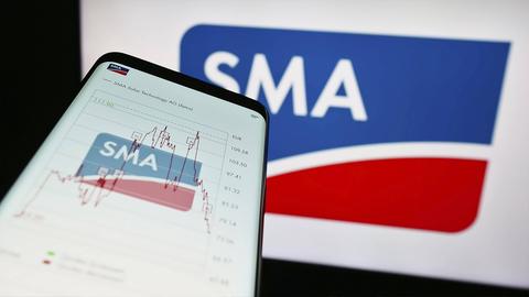 Smartphone in Nahaufnahme auf dessen Bildschirm ein Logo "SMA" und ein erste steigender und dann abfallender Aktionkurs zu sehen sind.