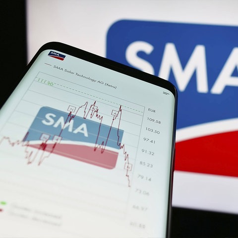 Smartphone in Nahaufnahme auf dessen Bildschirm ein Logo "SMA" und ein erste steigender und dann abfallender Aktionkurs zu sehen sind.