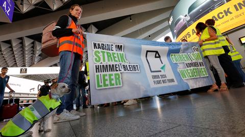 Menschen und ein Hund in Streik-Warnwesten in einem Flughafengebäude, auf einem Banner steht "Kabine im Streik, Himmel bleibt leer"