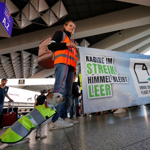 Menschen und ein Hund in Streik-Warnwesten in einem Flughafengebäude, auf einem Banner steht "Kabine im Streik, Himmel bleibt leer"