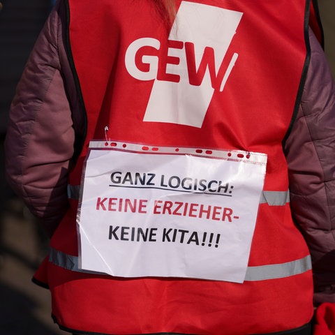 Eine Kundgebungsteilnehmerin trägt bei einem Streik die Aussage „Ganz logisch: Keine Erzieher- Keine Kita!!!“ auf dem Rücken über einer Weste der Gewerkschaft GEW.