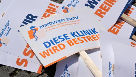 Einige Streiktafeln und Flyer mit den Aufschriften "Diese Klinik wird bestreikt" und "Marburger Bund" in orangfarbener und blauer Schrift liegen übereinander.