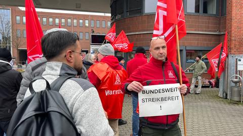 Ein Mitglied der IG Metall steht vor dem Haupttor in Baunatal. In den Händen hält er ein Schild. Darauf steht "Bundesweit streikbereit".