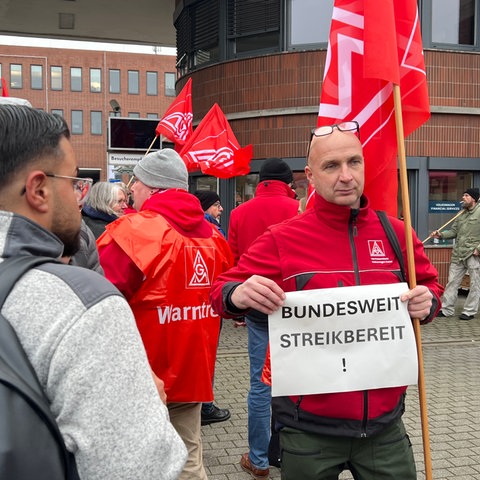 Ein Mitglied der IG Metall steht vor dem Haupttor in Baunatal. In den Händen hält er ein Schild. Darauf steht "Bundesweit streikbereit".