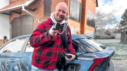 André Scheidler steht vor seinem E-Auto - mit dem Stecker in der Hand. Der 32-Jährige, Glatze, kurzer Vollbart, trägt einen rotkarierten Kapuzenpulli und lächelt in die Kamera.