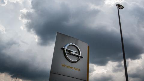 Opel-Symbol auf einer Stele vor dem Werk in Rüsselsheim - aus der Froschperspektive fotografiert. Darüber schwebt eine dunkle Wolke am Himmel.