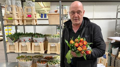 Ein Mann in schwarzer Kleidung hält in einer Lagerhalle einen Strauß Tulpen hoch.