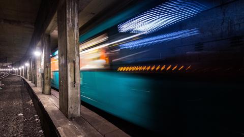 Foto: in einem Tunnel mit vielen Säulen (scharf) fährt eine leuchtende U-Bahn (unscharf).