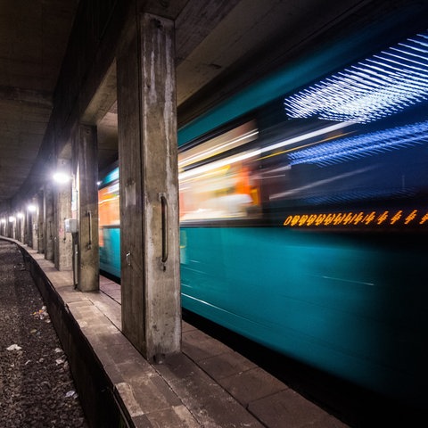 Foto: in einem Tunnel mit vielen Säulen (scharf) fährt eine leuchtende U-Bahn (unscharf).