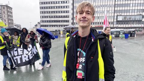 Ein junger Mann mit blonden Haaren steht auf einem großen Platz. Er trägt eine gelbe Warnweste und eine rote Trillerpfeife um den Hals. Im Hintergrund sind Demonstrierende zu sehen.