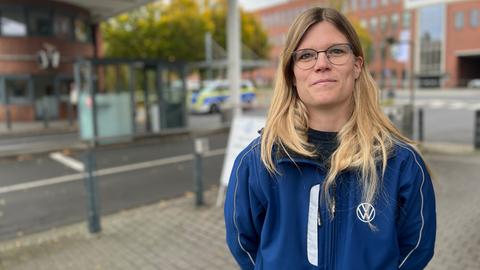 Eine Frau mit langen, blonden Haaren steht vor dem Haupttor bei VW in Baunatal. Sie trägt eine Brille und eine blaue Jacke mit VW-Logo.