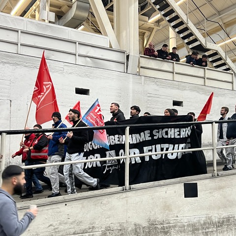 Mitgleider der Jugend der IG Metall kommt eine Rampe in die Halle runter. Sie tragen ein schwarzes Banner mit der Aufschrift "Übernahme sichern - for our future"