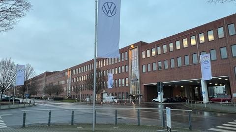 Blick auf das VW-Werk in Baunatal (Kassel). Das Werk ist eine langgezogene Fabrikhalle mit vielen Fenstern. Im Vordergrund stehen drei Fahnen mit dem VW-Logo (V über W in einem Kreis).