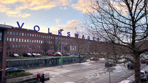 Volkswagenwerk Kassel am Standort Baunatal: auf dem Dach leuchten blaue Buchstaben als Wort "Volkswagen". Die Fassade aus rotem Stein hat unzählige Fenster. Davor parken Autos.