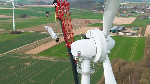 Foto aus der Vogelperspektive: Ein winzig aussehender Arbeiter sitzt auf der Spitze eines riesigen Windrades und greift nach einem schwebenden Rotorblatt, das frei an einem Kran hängt.