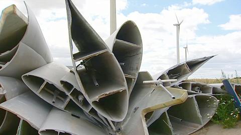 Im Bildvordergund einzelne Rotorblätter liegen auf einem Haufen am Boden. Im Hintergund unscharf Landschaft mit Windrädern in Funktion.