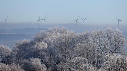 Im Vordergrund einige blätterlose Bäume mit Raureif an den Ästen und Zweigen. Im Hintergrund im Nebel sind einige Windkraftanlagen zu erkennen. 