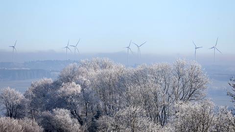 Im Vordergrund einige blätterlose Bäume mit Raureif an den Ästen und Zweigen. Im Hintergrund im Nebel sind einige Windkraftanlagen zu erkennen. 