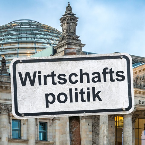 Ein weißes Hinweisschild mit der Aufschrift "Wirtschaftspolitik", befestigt mit Schrauben an einem Schilderpfahl, vor dem dem Berliner Reichstagsgebäude - es handelt sich u m eine Bildmontage